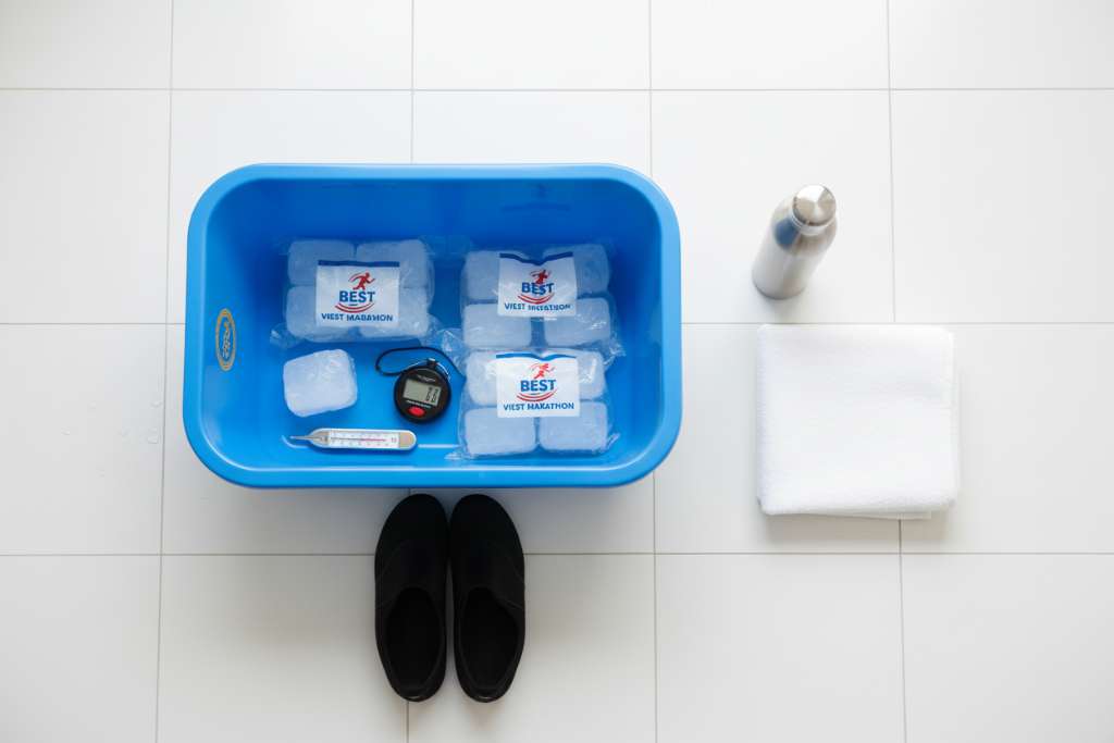 Flat lay trên nền sàn gạch trắng sáng gồm các dụng cụ ngâm đá tại nhà: thùng nhựa xanh dương 150 lít, 3 túi đá cây đóng gói, nhiệt kế nước analog, đồng hồ bấm giờ nhỏ, đôi giày neoprene đen, khăn bông trắng gấp gọn, bình giữ nhiệt inox. Góc chụp từ trên xuống (top-down), ánh sáng tự nhiên đều, phong cách minimal sạch sẽ.